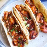 three tacos de cameron on a plate topped with cotija cheese and lettuce with limes in the background