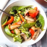 white bowl with pad pad thai which contains broccoli, snow peas, red bell peppers, mushrooms, carrot topped with sesame seeds