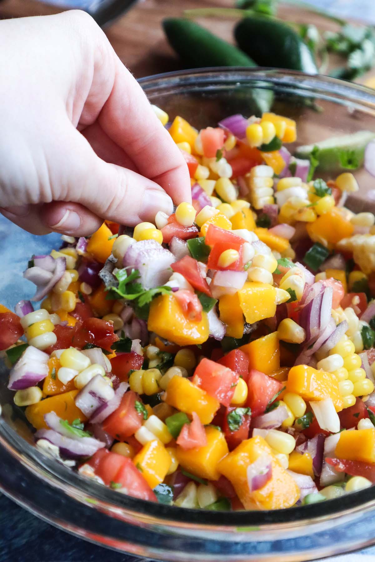 glass bowl with corn mango salsa with red onion, tomatoes, jalapeño, cilantro with a chip holding a scoop of the salsa.