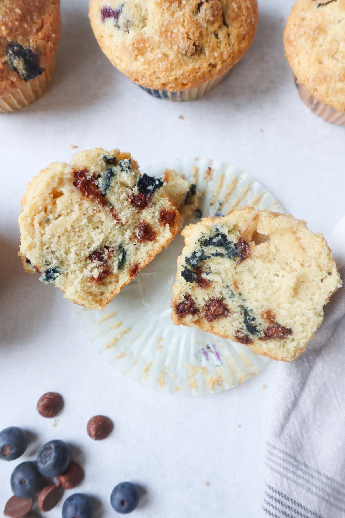 one blueberry chocolate chip muffin cut in half surrounded by other muffins and blueberries and chocolate chips