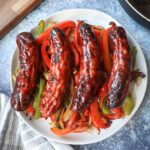 white plate with four chicken sausages on top of green and red peppers with onion