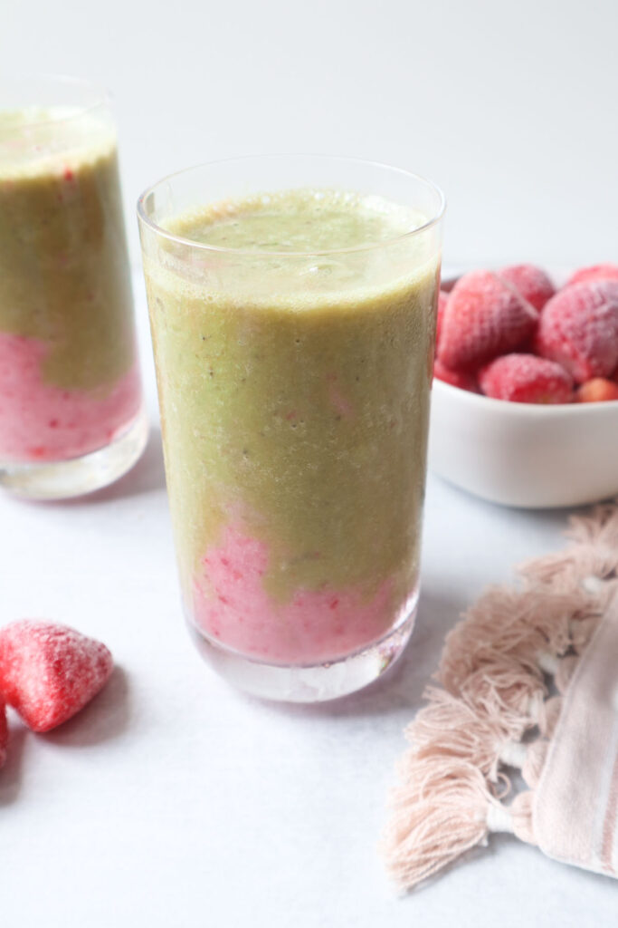 two glasses with strawberry matcha smoothie with a pink strawberry layer at the bottom and a green matcha layer on top with a bowl of strawberries behind the smoothie.