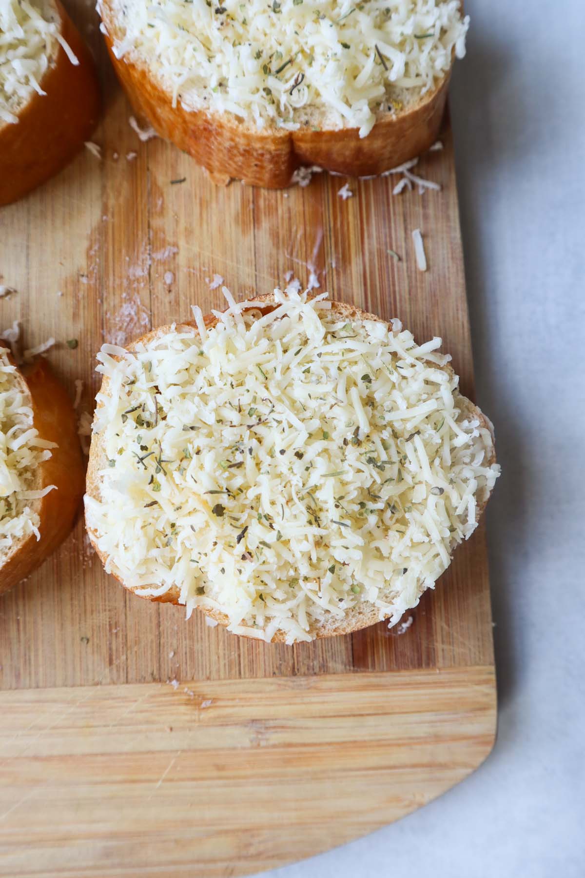 wooden cutting board with one slice of french bread topped with garlic herb butter and shredded cheese with a dash of italian seasoning