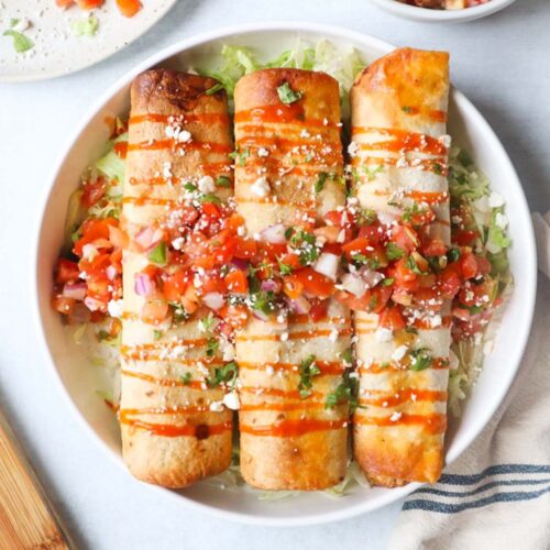 white plate with three chicken flautas on top of a bed of lettuce, topped with hot sauce, pico de gallo, and cotija cheese
