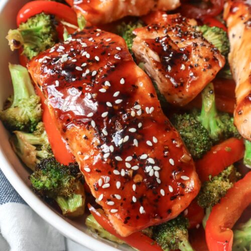 white plate with red bell pepper, broccoli, cooked salmon topped with sauce and sesame seeds
