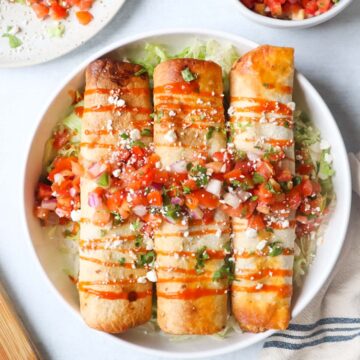 white plate with three air fry flautas on top of a bed of lettuce, topped with hot sauce, cotija cheese, and pico de gallo