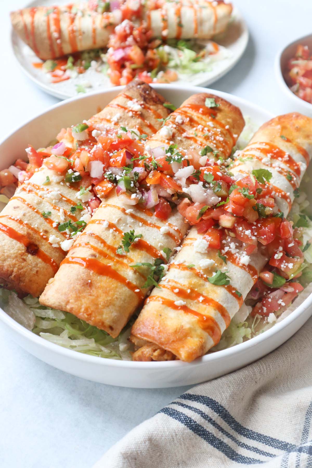 white plate with three air fry flautas on top of a bed of lettuce, topped with hot sauce, cotija cheese, and pico de gallo