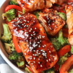 white bowl with red bell pepper, broccoli, cooked salmon topped with sauce and sesame seeds