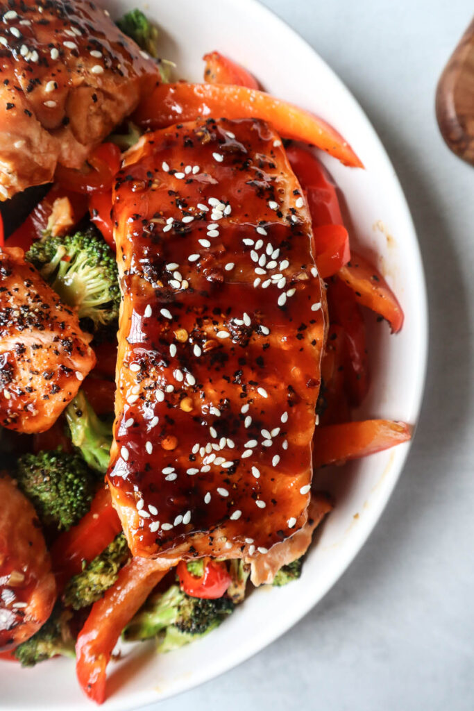 white bowl with red bell pepper, broccoli, cooked salmon topped with sauce and sesame seeds