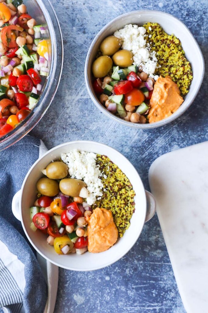 two white bowls with mediterranean power bowls topped with tomato cucumber salad, olives, feta, and hummus