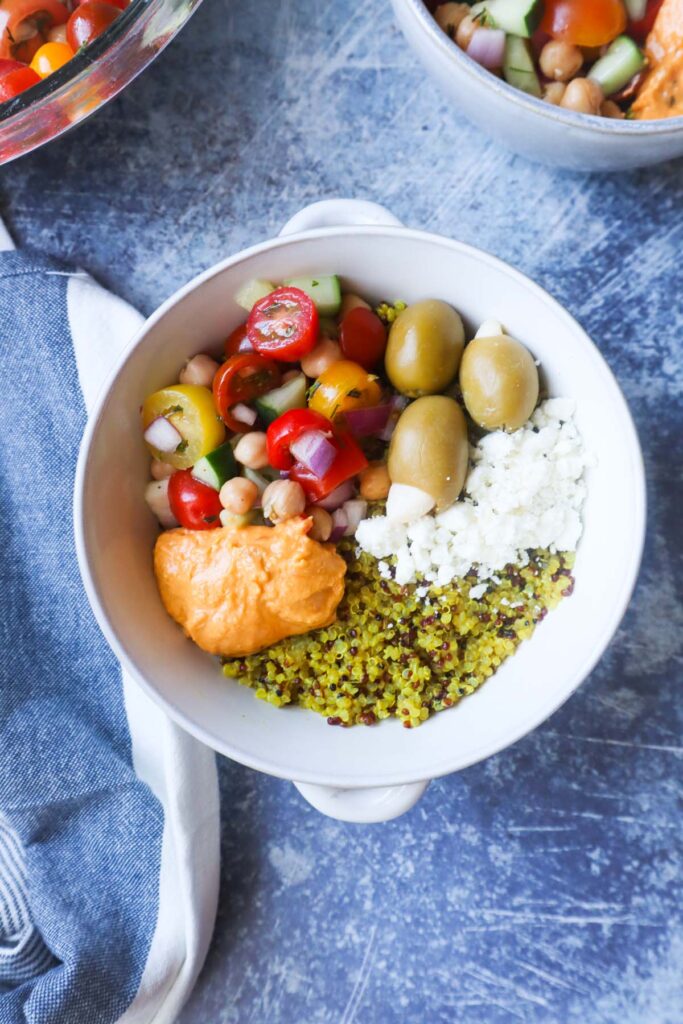 white bowl with quinoa, feta, olives, hummus, and tomato cucumber salad