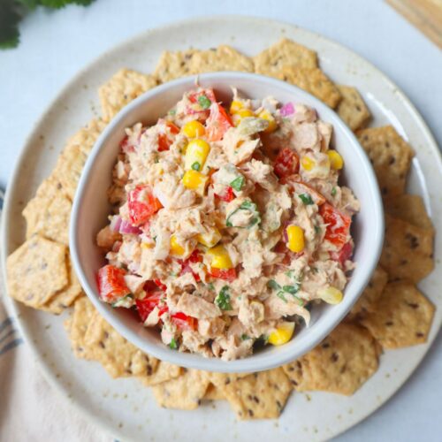 white bowl surrounded by crackers with tuna corn salad topped with herbs, red bell pepper, and onion