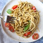 white plate with spaghetti, pesto, grape tomatoes, and two slices of basil