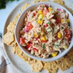 white bowl surrounded by crackers with tuna corn salad topped with herbs, red bell pepper, and onion