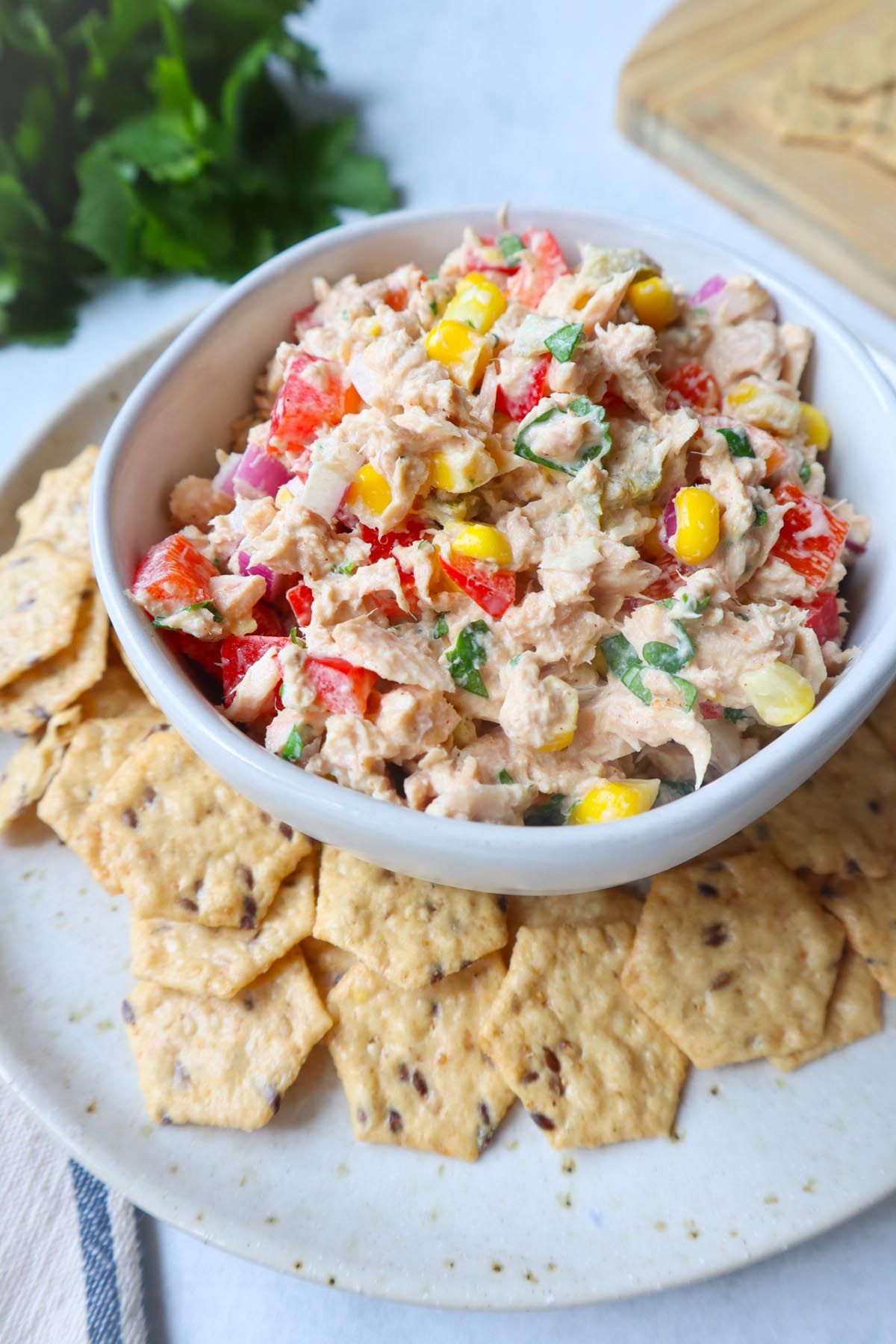 white bowl surrounded by crackers with tuna corn salad topped with herbs, red bell pepper, and onion
