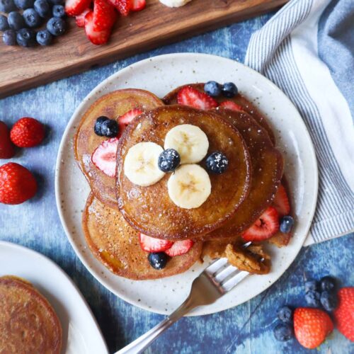 white plate with pumpkin pancakes topped with banana, blueberries, and strawberries, and maple syrup