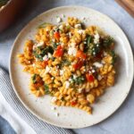 white plate with pasta tossed in a pumpkin feta sauce topped with red bell pepper and spinach