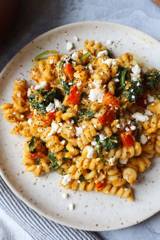 white plate with pasta tossed in a pumpkin feta sauce topped with red bell pepper and spinach