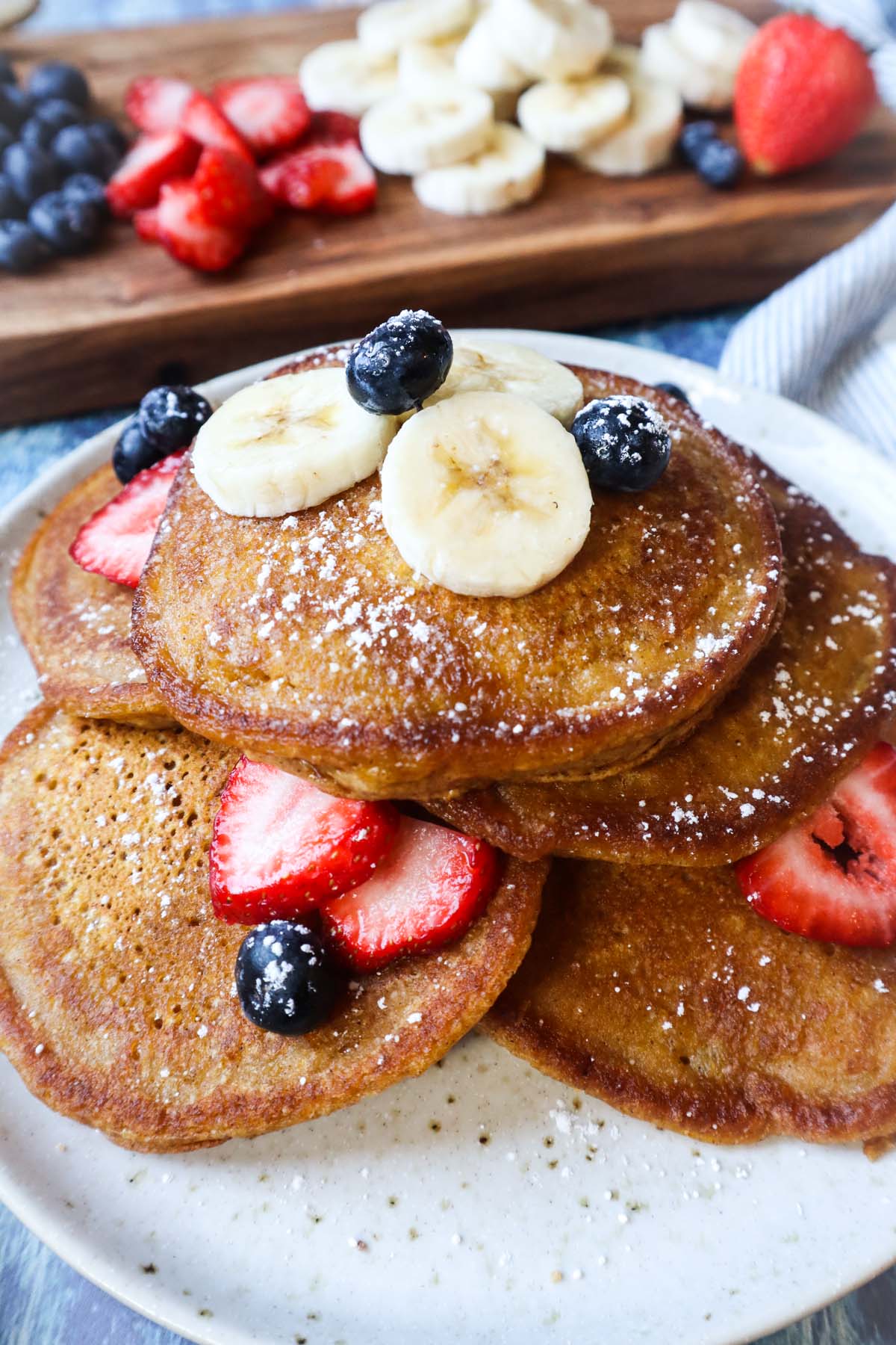 white plate with pumpkin pancakes topped with banana, blueberries, and strawberries, and powdered sugar