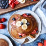 white plate with pumpkin pancakes topped with banana, blueberries, and strawberries, and maple syrup