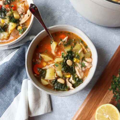 white bowl with a spoon with chicken soup with beans, kale, potatoes, carrots, and more.