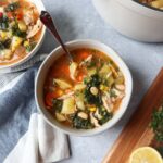 white bowl with a spoon with chicken soup with beans, kale, potatoes, carrots, and more.