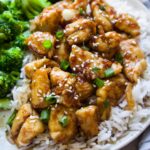 white plate with chicken over top of white rice and a side of broccoli topped with sesame seeds and green onion.