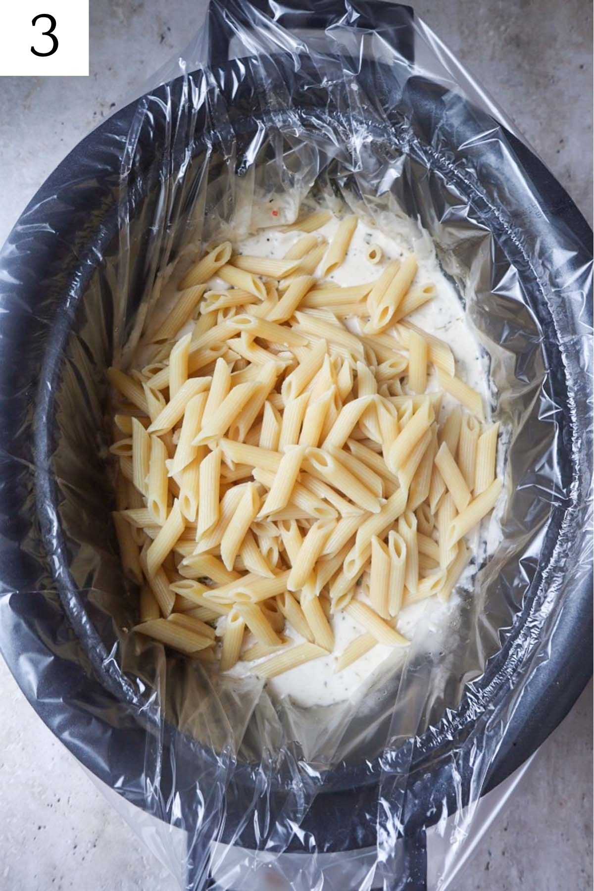 crock pot with broccoli alfredo topped with cooked penne.