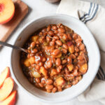 white bowl with peach baked beans surrounded by fresh peaches.