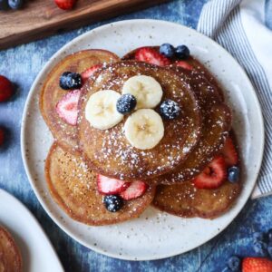 pumpkin pancakes on a plate topped with fresh fruit and powdered sugar.