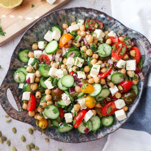 bowl with cucumber chickpea feta salad topped with various herbs and tossed in the dressing.