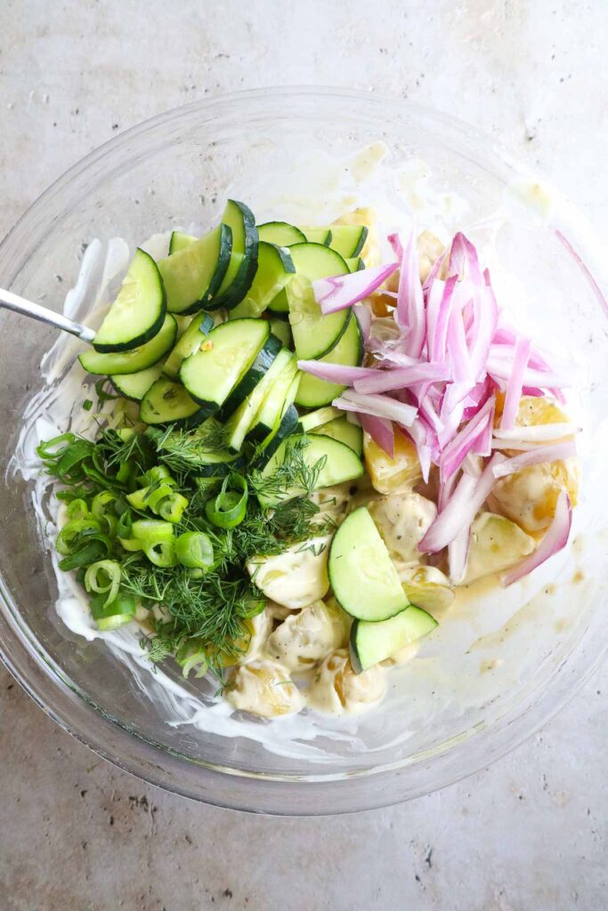 glass bowl with cucumber, red onion, and other ingredients mixed into the potatoes and dressing.